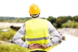 Construction worker in a yellow safety vest and hard hat holding his lower back in pain at a construction site.